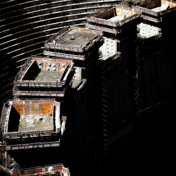A large inşaat sahası with several elevated, rectangular concrete structures surrounded by scaffolding. Workers in safety gear are visible on the platforms. The background shows curved, tiered earthworks suggesting a large excavation or dam. There are shadows that cast a dramatic contrast over the scene.